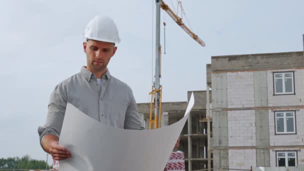 Un arquitecto en un casco, la gestión de un proyecto, de pie con dibujos en un sitio de construcción. El concepto de construir un edificio de varios pisos — Vídeo de stock