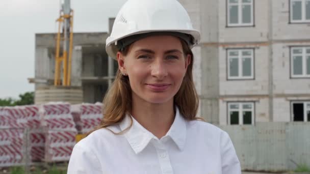 Portret van een vrolijke vrouwelijke architect in een witte helm op een bouwplaats tegen de achtergrond van een huis in aanbouw — Stockvideo