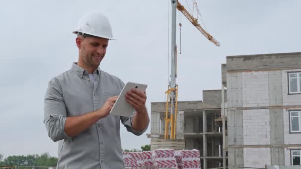 Ingénieur de construction dans un casque blanc avec une grue jaune en arrière-plan utilise une tablette — Video