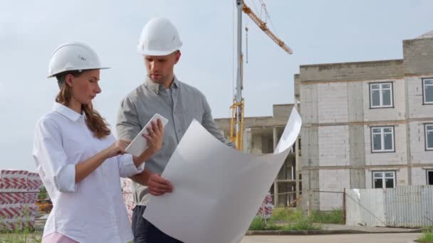 Γυναίκα και ένας άνθρωπος στην κατασκευή λευκά κράνη με σχέδια στα χέρια τους στο παρασκήνιο ενός κτιρίου υπό κατασκευή. Προγραμματισμός κατασκευών. — Αρχείο Βίντεο