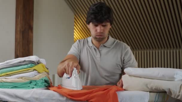 Niezadowolony człowiek wykonuje prace domowe i prasuje ubrania. Zmęczony człowiek chwyta się za głowę. — Wideo stockowe