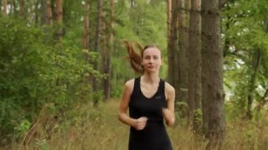 Genç bir kadın orman yolunda çam ormanının arka planında koşuyor.