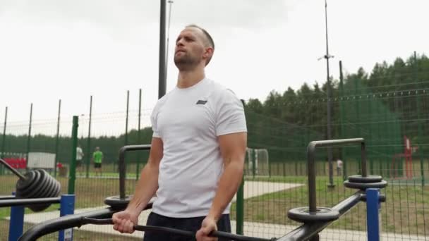 El hombre está involucrado en un simulador en un campo de deportes en el parque. Un hombre sacude sus bíceps en un simulador especial — Vídeo de stock