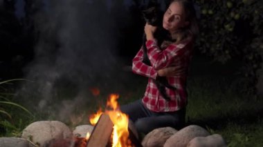 Kadın bir kedi tutuyor ve akşam kamp ateşinin yanında oturuyor.