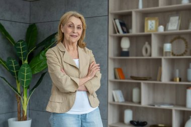 70 'lerin olgun iş kadını, odada duruyor. Yaşlı, orta yaşlı, stil sahibi, kollarını kavuşturup kameraya bakan kadın portresi.