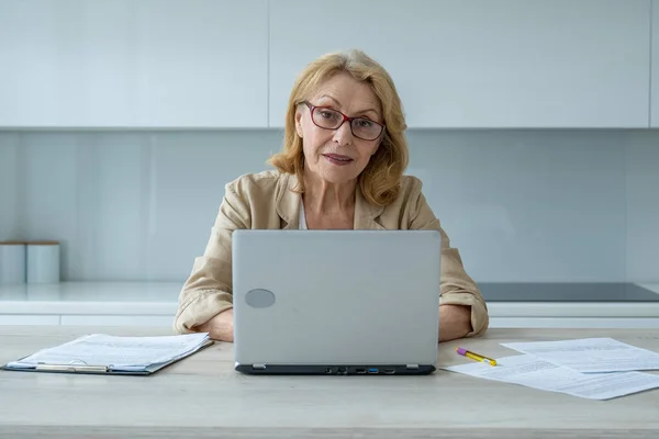 Feliz anciana con gafas y mirando a la cámara en casa. Una anciana exitosa trabaja en casa en una computadora — Foto de Stock
