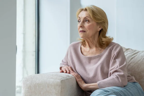 Uma mulher idosa pensativa está sentada no sofá na sala de estar, olhando pela janela para a distância, pensando ou pensando — Fotografia de Stock