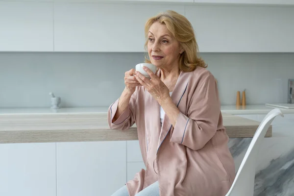 Atractiva anciana disfrutando de una taza de café en su cocina pensando en agradecimiento. — Foto de Stock