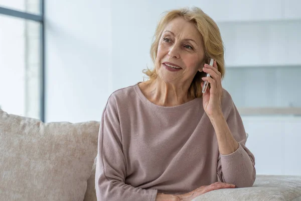 Mujer anciana sonriente hablando en un teléfono móvil en casa, relajándose en el sofá en la sala de estar — Foto de Stock