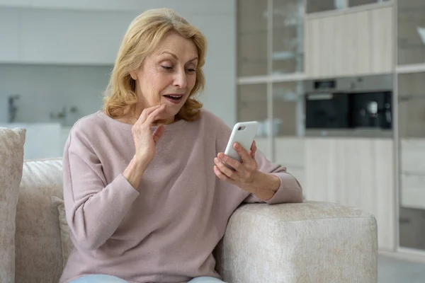 Una anciana entusiasta usando un smartphone, impresionada por las redes sociales, sentada en el sofá de la sala de estar — Foto de Stock