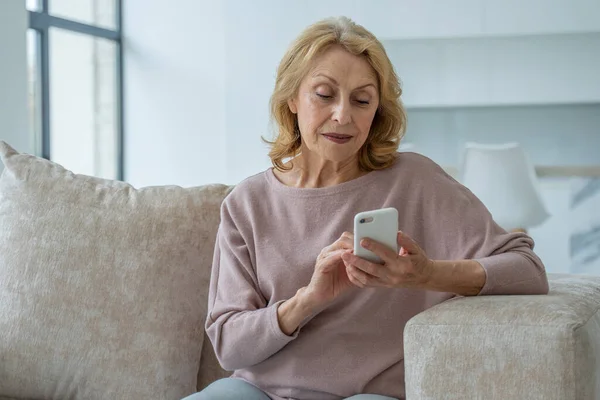 Una abuela encantadora sentada en el sofá en la sala de estar de la casa, haciendo compras en línea con un teléfono inteligente en sus manos — Foto de Stock