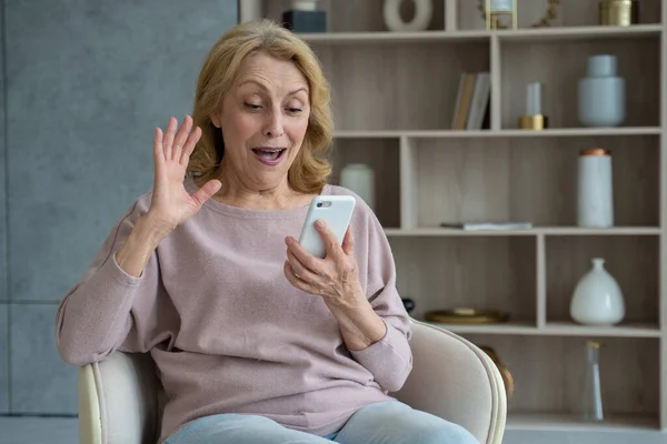 Una abuela jubilada jubilada y encantada celebra la victoria en el juego en su teléfono móvil. Una anciana feliz está sentada en una silla y leyendo e - mail, emocionada por un descuento en una venta en — Foto de Stock