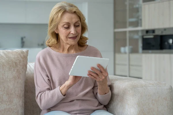 Mujer de mediana edad sentada en el sofá de la sala de estar, divirtiéndose con una tableta, utilizando el concepto del dispositivo — Foto de Stock