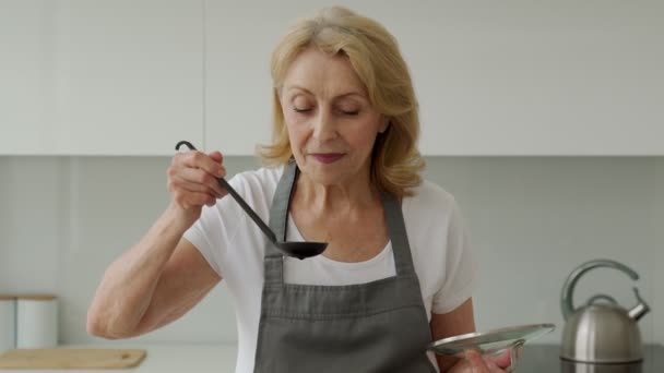 Wanita tua yang lucu memegang sendok-sendok untuk memasak sambil makan sup di rumah — Stok Video