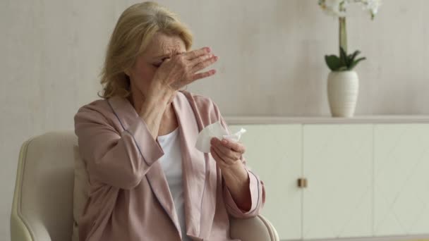 Egy beteg idős nő tüsszentett papírszalvétát használva az orrára, folyt az orra és köhögött egy széken a nappaliban. — Stock videók