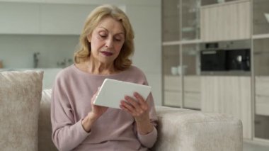 Orta yaşlı bir kadın oturma odasındaki koltukta oturuyor, tabletle eğleniyor, cihaz konseptini kullanıyor.