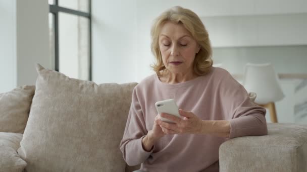 Uma avó encantadora sentada no sofá na sala de estar da casa, fazendo compras on-line com um smartphone em suas mãos — Vídeo de Stock