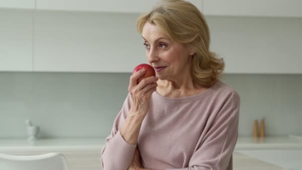 Una hermosa anciana sonriente sosteniendo una manzana roja en sus manos, de pie en su cocina — Vídeo de stock