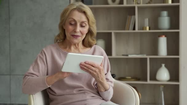 Una donna anziana seduta su una poltrona e con un tablet digitale seduta su una poltrona nel suo salotto — Video Stock