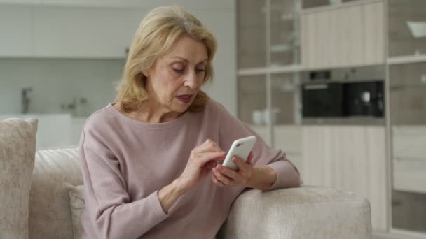 Nadšená stařena používající smartphone, ohromená sociálními sítěmi, sedící na gauči v obývacím pokoji — Stock video