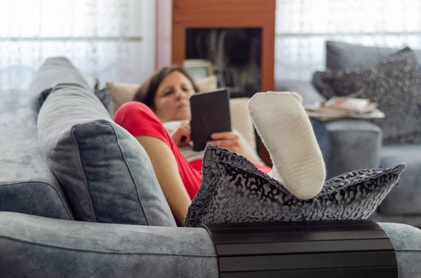 The middle aged woman with broken leg  is lying on the sofa and she is reading  her messages at home,selective focus