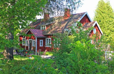 Geleneksel, kırmızı ahşap bir ev ve güneşli yaz akşamında yeşil yaprakların arkasındaki avlu. Margreteberg, Espoo, Finlandiya. 