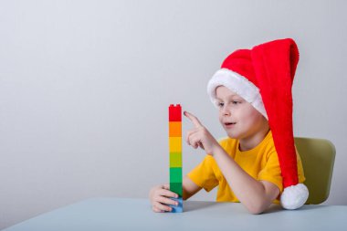 Çocuk Noel Baba şapkasıyla masada otururken parmaklarını sayıyor. Masanın üzerinde renkli şekiller sıra halinde bloklar var.