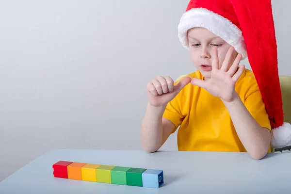 Rapaz a contar com os dedos sentados numa mesa de chapéu de Pai Natal. sobre a mesa formas coloridas blocos em uma linha — Fotografia de Stock