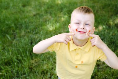 Ketçaplı şirin, kirli çocuk gözlerini kapattı, gülümsedi ve parmaklarını yanaklarından tuttu. Açık havada yaz. Yüksek kalite fotoğraf