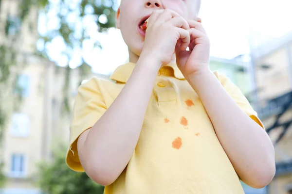 男の子はフランスのホットドッグを噛み ケチャップの汚れで服を汚します 公園で 衣類の汚れをきれいにするというコンセプト 高品質の写真 — ストック写真