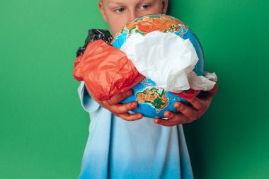 Çocuk, yeşil arka planda, ortasından plastik tek kullanımlık paketler ile Dünya 'yı tutuyor. Dünyayı kurtar, yaratıcı, çevre kirliliği Dünya Günü konseptimiz.