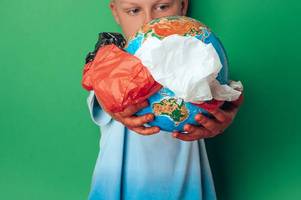 Niño sosteniendo el planeta Tierra con paquetes desechables de plástico del medio sobre un fondo verde. Salvar el mundo, creativo, la contaminación del medio ambiente nuestro concepto del Día Mundial de la Tierra. — Foto de Stock