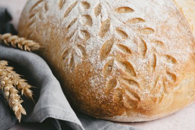  Bir somun zanaatkar ekmeği, gri bir peçetenin üzerinde duruyor. Kabuğunda karmaşık yaprak desenleri var. Etrafını saran altın buğday sapları kırsal çekiciliğini vurguluyor. Yüksek kalite fotoğraf