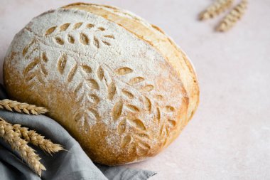  Bir somun zanaatkar ekmeği, gri bir peçetenin üzerinde duruyor. Kabuğunda karmaşık yaprak desenleri var. Etrafını saran altın buğday sapları kırsal çekiciliğini vurguluyor. Yüksek kalite fotoğraf
