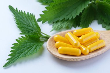 Tahta bir kaşıkta sarı bitkisel kapsüller temiz beyaz bir yüzey üzerinde canlı yeşil yaprakların yanında sergileniyordu. Doğal malzemeler, alternatif tıp, naturopati, beslenme takviyesi..