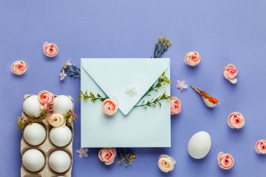 Paskalya tebrik kartı, yumurta tepsisinde el yapımı şirin yumurtalar, kraft mavisi zarf ve leylak arka planda gül çiçekleri. Düz konsept, kopyalama alanı olan üst görünüm.