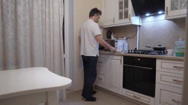 Kafkasyalı bir adam sıcak mutfakta yemek ısıtıyor, ev yemeği pişiriyor, mikrodalgaya sıcak LED ışıklandırma, gündelik tişört, modern dolaplar ve sergilenen tezgah, rahat akşam rutini.