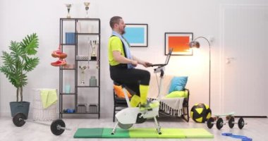 Formda bir adam evde bisikletle antrenman yapıyor ve bilgisayarla çalışıyor. Sporcu, sporcu çevrimiçi video eğitimi kullanarak evde çalışıyor.