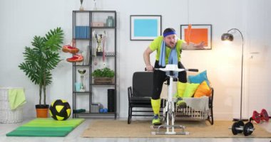 Sabit bisiklete binen genç bir sporcu evde egzersiz yapıyor. Fitness parodisi. Mizah. Spor komedisi