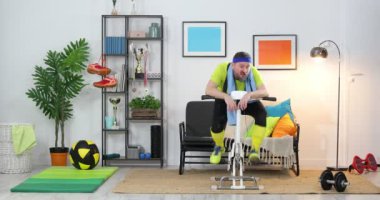 Sabit bisiklete binen genç bir sporcu evde egzersiz yapıyor. Fitness parodisi. Mizah. Spor komedisi