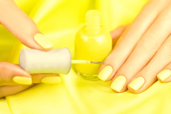 Female hands with yellow manicure on a yellow silk background. Woman painting her nails with yellow nail varnish. Close up