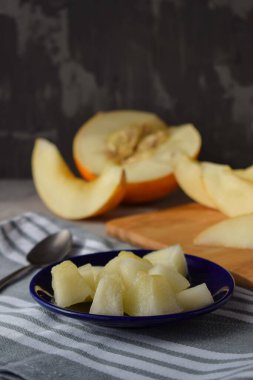 Dilimlenmiş kavun parçaları küçük mavi bir tabakta, gri bir bezin üzerindeki çay kaşığının yanında yarım kavun ve dilimlerinin arka planında.