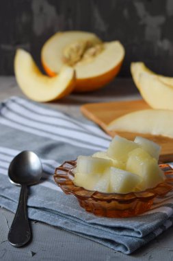 Dilimlenmiş kavun parçaları küçük cam bir tabakta, gri bir bezin üzerindeki çay kaşığının yanında yarım kavun ve dilimlerinin arka planında.