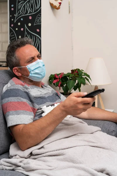Male model covered with blanket wearing protective disposable medical or surgical face mask watching tv holding remote control from sofa at home sick having sars influenza symptoms