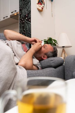 Sinüzit soğuk algınlığı belirtileri gösteren hasta yetişkin erkek modeli evdeki kanepede dinlenen peçeteyle burnunu siliyor ve masada salgın konsepti olarak bir fincan çay var.