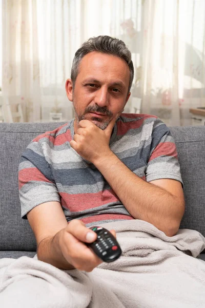 Ill man during pandemic at home watching tv from couch with curious expression covered by blanket holding remote controller