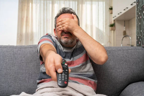 Adult man making scared afraid gesture covering eyes using hands watching horror movie on tv changing channel with remote control as at home pandemic concept