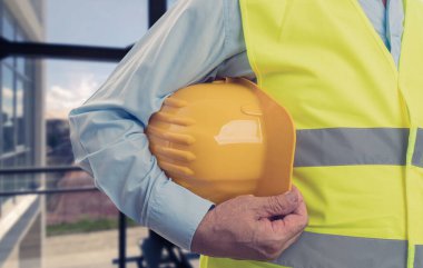 Flüoresan iş yeleği giyen yaşlı erkek müteahhitin yakın çekimi. Elinde düz şehir arka planına sahip sarı güvenlik başlığı var.