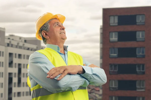 Portrait of old male constructor wearing fluorescent vest and yellow safety hardhat with arms crossed on building background