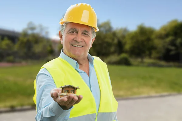 Old male builder in work attire smiling presenting miniature wooden house building concept park nature background